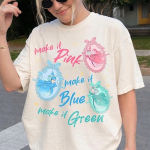 Puede incluir: Camiseta color crema con el texto "make it Pink, Blue, Green" e ilustraciones de hadas en rosa, azul y verde. El diseño incluye marcos ovalados decorativos y lazos, creando una estética caprichosa.