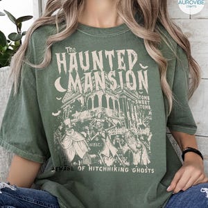 May include: Green t-shirt featuring a vintage-style graphic of "The Haunted Mansion" with a spooky house, bats, and ghostly figures. The text includes "Tomb Sweet Tomb" and "Beware of Hitchhiking Ghosts."