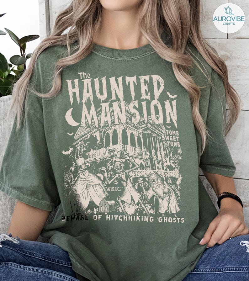 May include: Green t-shirt featuring a vintage-style graphic of "The Haunted Mansion" with a spooky house, bats, and ghostly figures. The text includes "Tomb Sweet Tomb" and "Beware of Hitchhiking Ghosts."
