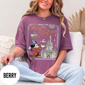 May include: Berry-colored t-shirt featuring a graphic of Mickey Mouse in a wizard outfit, the Magic Kingdom logo, and a castle. The design includes a tram, a ship, and stars. The shirt is a short-sleeved, oversized fit.