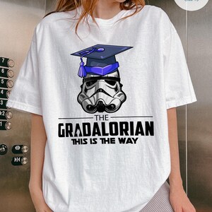 Pu&ograve; includere: T-shirt bianco con grafica di un casco da Stormtrooper con cappello di laurea. Il testo recita "THE GRADALORIAN THIS IS THE WAY" in nero. La maglietta ha una vestibilit&agrave; ampia.