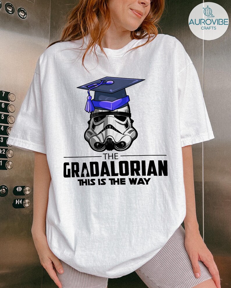 Pu&ograve; includere: T-shirt bianco con grafica di un casco da Stormtrooper con cappello di laurea. Il testo recita "THE GRADALORIAN THIS IS THE WAY" in nero. La maglietta ha una vestibilit&agrave; ampia.