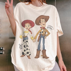 Może przedstawiać: Kremowy t-shirt z kresk&oacute;wkową ilustracją Jessie i Woody'ego z Toy Story. Jessie ma na sobie czerwony kapelusz, białą koszulę i spodnie w krowie łaty. Woody ma kapelusz kowbojski, kamizelkę i niebieskie dżinsy. Na koszulce widnieje napis "Woody".