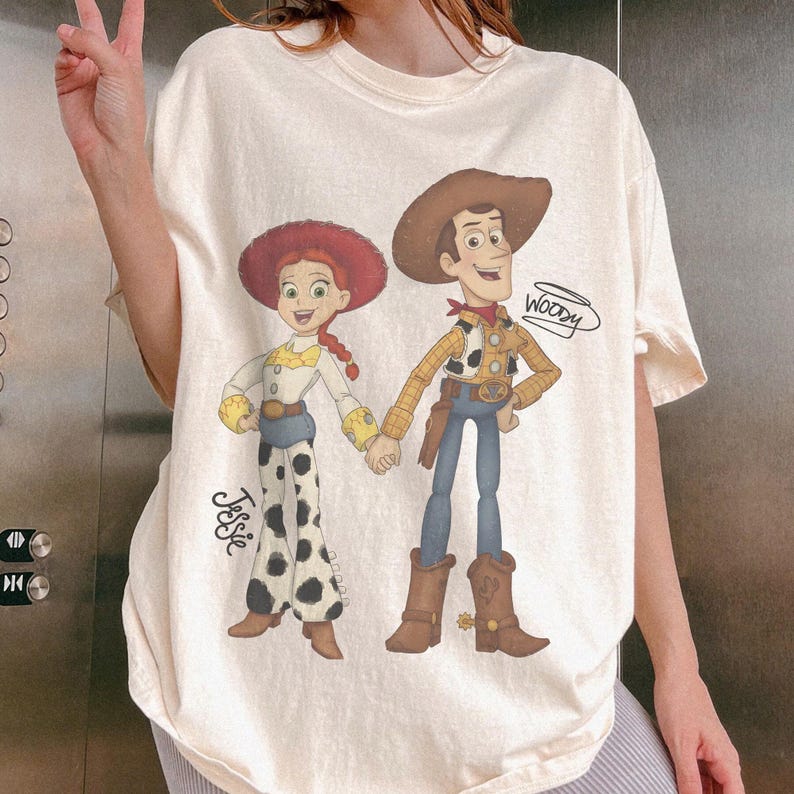 Może przedstawiać: Kremowy t-shirt z kresk&oacute;wkową ilustracją Jessie i Woody'ego z Toy Story. Jessie ma na sobie czerwony kapelusz, białą koszulę i spodnie w krowie łaty. Woody ma kapelusz kowbojski, kamizelkę i niebieskie dżinsy. Na koszulce widnieje napis "Woody".
