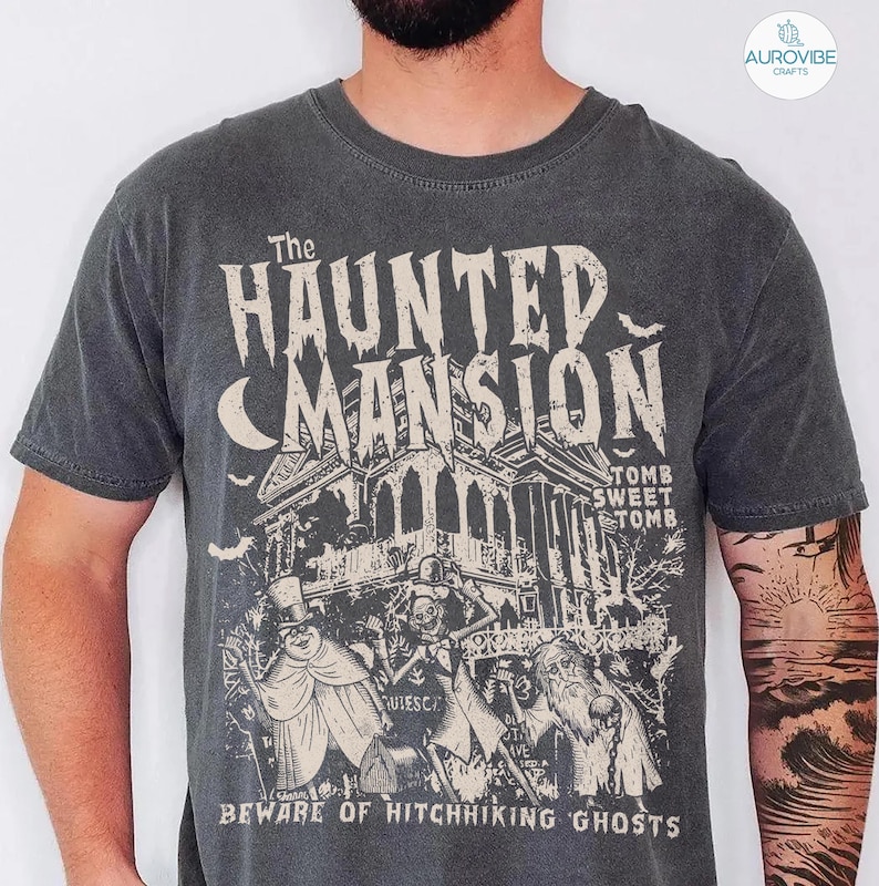 May include: Dark gray t-shirt featuring a vintage-style graphic of The Haunted Mansion. The design includes the text "The Haunted Mansion," a spooky mansion illustration, and the phrase "Beware of Hitchhiking Ghosts."