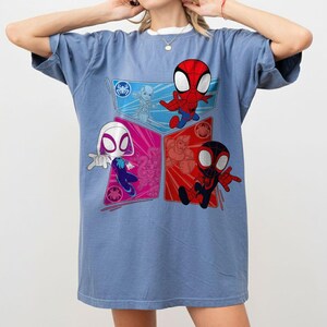 May include: A light blue t-shirt featuring a colorful graphic of Spider-Man characters. The design includes Spider-Man, Spider-Gwen, and Miles Morales in a cartoon style. The shirt has a crew neck and short sleeves.