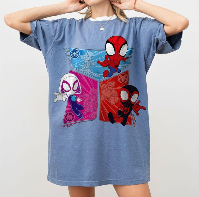 May include: A light blue t-shirt featuring a colorful graphic of Spider-Man characters. The design includes Spider-Man, Spider-Gwen, and Miles Morales in a cartoon style. The shirt has a crew neck and short sleeves.