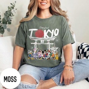 Puede incluir: Camiseta verde musgo con el texto "Disneyland TOKYO" sobre un gr&aacute;fico de una puerta japonesa y personajes de dibujos animados. La camiseta presenta un gr&aacute;fico redondo rojo y una fila de personajes de dibujos animados.