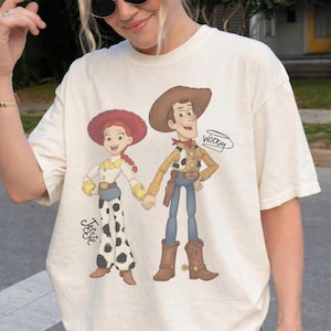 Może przedstawiać: Kremowy t-shirt z kresk&oacute;wkową ilustracją Woody'ego i Jessie z Toy Story, trzymających się za ręce. Woody ma na sobie kapelusz kowbojski, kamizelkę i niebieskie dżinsy. Jessie ma czerwony kapelusz, białą koszulę i spodnie w krowie łaty. Na koszulce widnieje napis "Woody" i "Jessie".