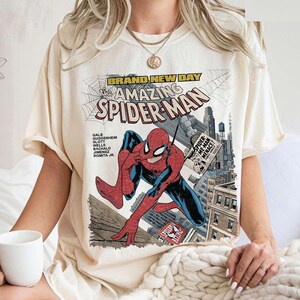 May include: Cream-colored t-shirt featuring a comic book cover of Spider-Man. The graphic includes the text "BRAND NEW DAY" and "The Amazing Spider-Man." The superhero is depicted in a dynamic pose with a cityscape backdrop.