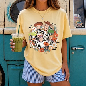 May include: A pale yellow short-sleeve t-shirt featuring a graphic print of Toy Story characters dressed in skeleton costumes, surrounded by pumpkins, flowers, spiderwebs, and bats. The shirt is paired with blue and white striped shorts.