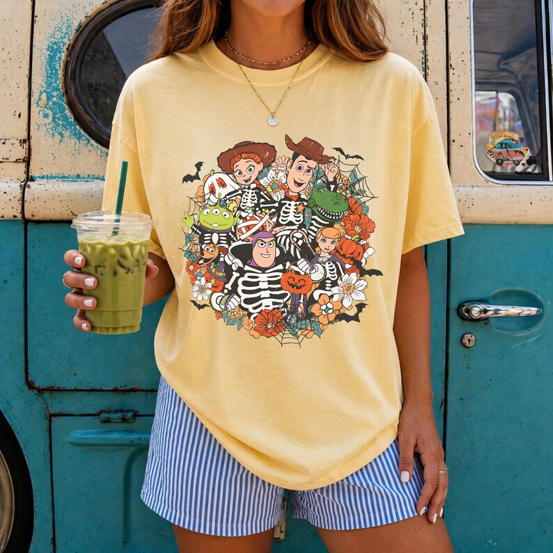 May include: A pale yellow short-sleeve t-shirt featuring a graphic print of Toy Story characters dressed in skeleton costumes, surrounded by pumpkins, flowers, spiderwebs, and bats. The shirt is paired with blue and white striped shorts.