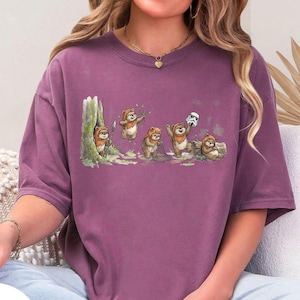 May include: A purple t-shirt featuring a whimsical illustration of Ewoks in a forest setting. The design includes several Ewoks interacting with each other, one holding a Stormtrooper helmet. The artwork is detailed and colorful.