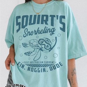 May include: A light blue t-shirt with the words "SQUIRT'S Snorkeling" and a cartoon turtle wearing a snorkel mask. The shirt also has the phrases "-FAST AUSTRALIAN CURRENT- FIN, NOGGIN, DUDE" printed on it. The shirt is a comfort color in chalky mint.