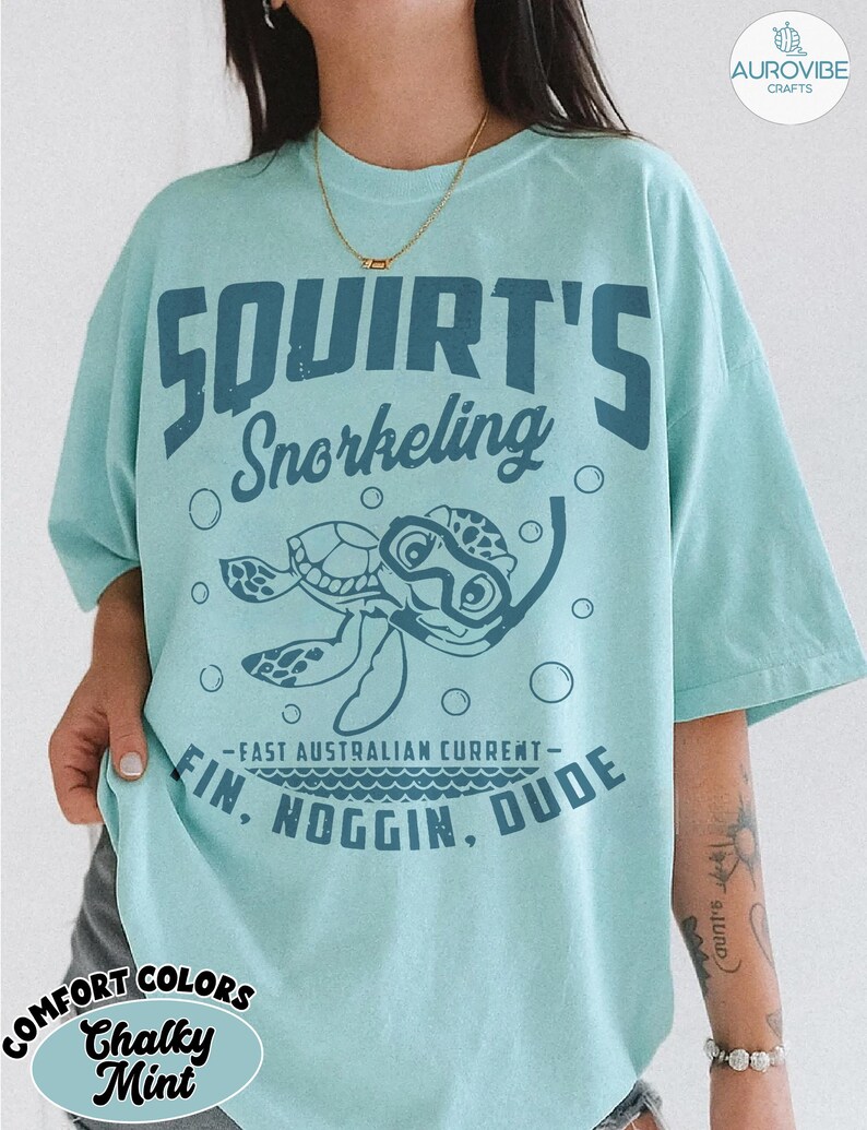 May include: A light blue t-shirt with the words "SQUIRT'S Snorkeling" and a cartoon turtle wearing a snorkel mask. The shirt also has the phrases "-FAST AUSTRALIAN CURRENT- FIN, NOGGIN, DUDE" printed on it. The shirt is a comfort color in chalky mint.
