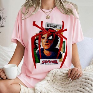 Op de afbeelding: Lichtroze T-shirt met een Spider-Man-afbeelding. Het ontwerp bevat een portret van het personage, een rood spinnenwebframe en de tekst "SPIDER-MAN BRAND NEW DAY". Extra tekst omvat "WITH GREAT POWER COMES GREAT RESPONSIBILITY" en "DAILY BUGLE".