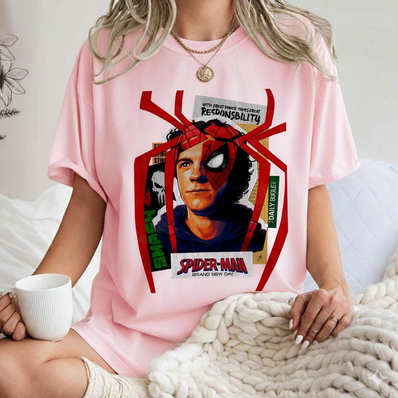 Op de afbeelding: Lichtroze T-shirt met een Spider-Man-afbeelding. Het ontwerp bevat een portret van het personage, een rood spinnenwebframe en de tekst "SPIDER-MAN BRAND NEW DAY". Extra tekst omvat "WITH GREAT POWER COMES GREAT RESPONSIBILITY" en "DAILY BUGLE".