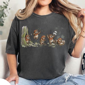 May include: Dark gray t-shirt featuring a Star Wars-themed illustration. The graphic depicts a series of Ewoks in various poses, including one holding a spear, another jumping, and others interacting with a Stormtrooper helmet. The design is set against a forest backdrop.