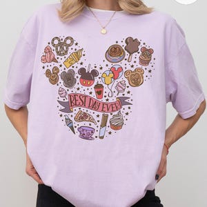 May include: A light purple t-shirt featuring a graphic print arranged in a Mickey Mouse head shape. The design includes various food items, balloons, and a banner with the text BEST DAY EVER. The shirt has a crew neckline and short sleeves.