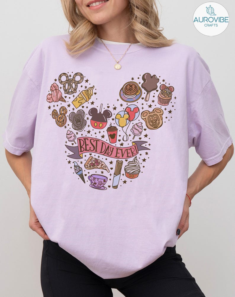 May include: A light purple t-shirt featuring a graphic print arranged in a Mickey Mouse head shape. The design includes various food items, balloons, and a banner with the text BEST DAY EVER. The shirt has a crew neckline and short sleeves.