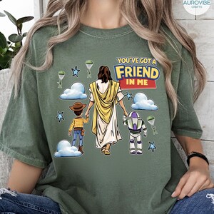 May include: Olive green t-shirt featuring a graphic of Jesus holding hands with two cartoon characters, with the text "YOU'VE GOT A FRIEND IN ME". The design includes clouds, stars, and parachutes.