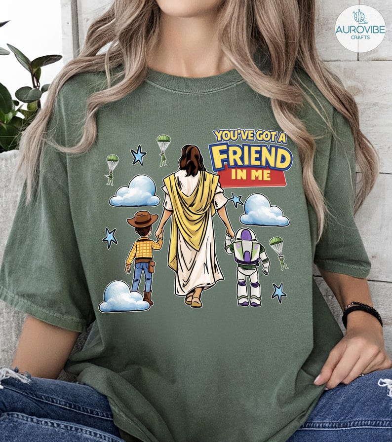 May include: Olive green t-shirt featuring a graphic of Jesus holding hands with two cartoon characters, with the text "YOU'VE GOT A FRIEND IN ME". The design includes clouds, stars, and parachutes.