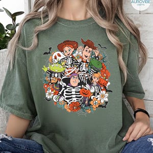 May include: An olive green t-shirt featuring a graphic print of animated characters dressed in skeleton costumes for Halloween. The design includes pumpkins, spiderwebs, and bats surrounding the characters, with vibrant orange and white accents.