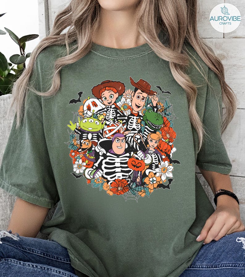 May include: An olive green t-shirt featuring a graphic print of animated characters dressed in skeleton costumes for Halloween. The design includes pumpkins, spiderwebs, and bats surrounding the characters, with vibrant orange and white accents.