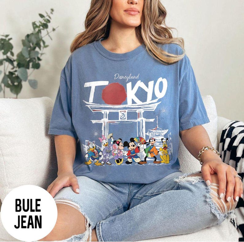 Puede incluir: Una camiseta azul con la palabra "TOKYO" en blanco, un c&iacute;rculo rojo y un dise&ntilde;o de puerta japonesa. Debajo del texto hay personajes de dibujos animados como Mickey Mouse, Minnie Mouse, Donald Duck y Goofy. La palabra "Disneyland" est&aacute; encima de "TOKYO".