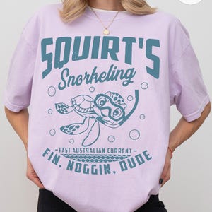 May include: A light purple t-shirt with the words "Squirt's Snorkeling" in teal. Below the text is a cartoon turtle wearing a snorkel. The shirt also has the text "Fin, Noggin, Dude" and "Fast Australian Current".