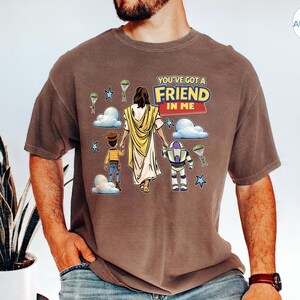 May include: Brown t-shirt with a graphic featuring the text "YOU'VE GOT A FRIEND IN ME". The graphic includes cartoon illustrations of a figure in a yellow robe, a cowboy, and a space ranger. The shirt is worn by a person.