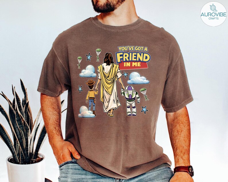 May include: Brown t-shirt with a graphic featuring the text "YOU'VE GOT A FRIEND IN ME". The graphic includes cartoon illustrations of a figure in a yellow robe, a cowboy, and a space ranger. The shirt is worn by a person.