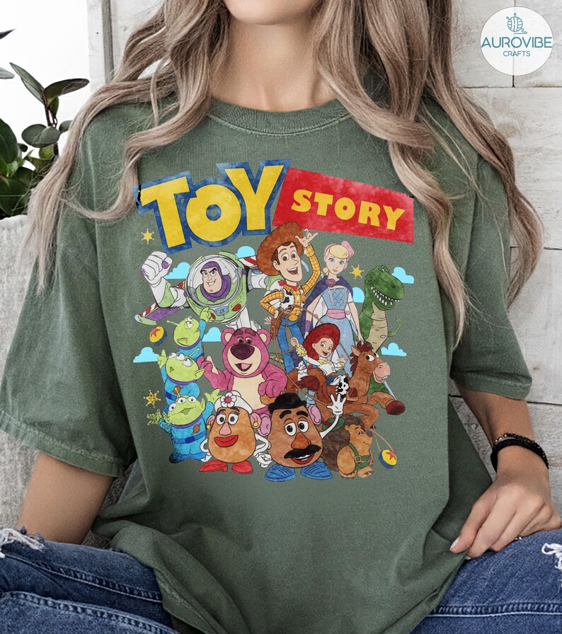May include: Green t-shirt featuring the words "TOY STORY" in yellow and red, with a graphic of various characters from the animated film. The design includes Woody, Buzz Lightyear, and other beloved characters.