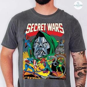 May include: Dark gray t-shirt featuring a comic book illustration with the text "SECRET WARS." The graphic depicts Spider-Man, Captain America, Iron Man, and Doctor Doom in a battle scene. The shirt has a vintage, distressed look.