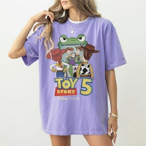 K&ouml;nnte beinhalten: Ein lila T-Shirt mit einem Grafikdesign aus dem Film Toy Story 5. Das Design zeigt Charaktere wie Woody und Buzz Lightyear, mit dem Filmtitel in einer farbenfrohen Schriftart. Das Disney Pixar-Logo befindet sich unten.