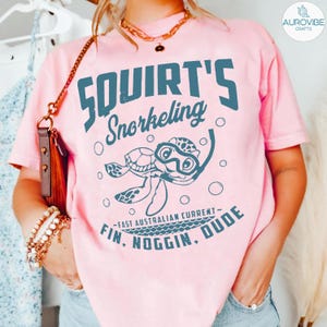 May include: Pink t-shirt with a teal graphic of a cartoon turtle wearing a snorkel and the words "Squirt's Snorkeling". The text "Fin, Noggin, Dude" is also printed on the shirt. The shirt is a light pink color.