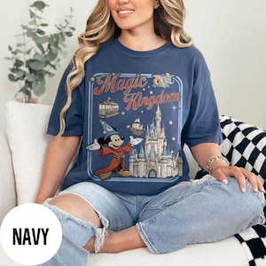 May include: Navy blue t-shirt featuring a graphic of Mickey Mouse in a sorcerer's outfit in front of a castle, with the words "Magic Kingdom". The design includes a boat, a carousel, and stars.