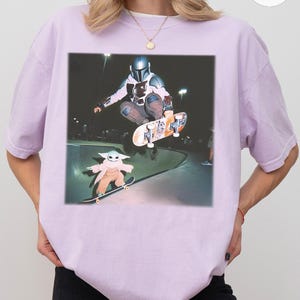 Puede incluir: Una camiseta lila claro con un gr&aacute;fico de un skater con casco mandaloriano en el aire, con un personaje de Baby Yoda patinando debajo. La imagen est&aacute; ambientada en un skate park.