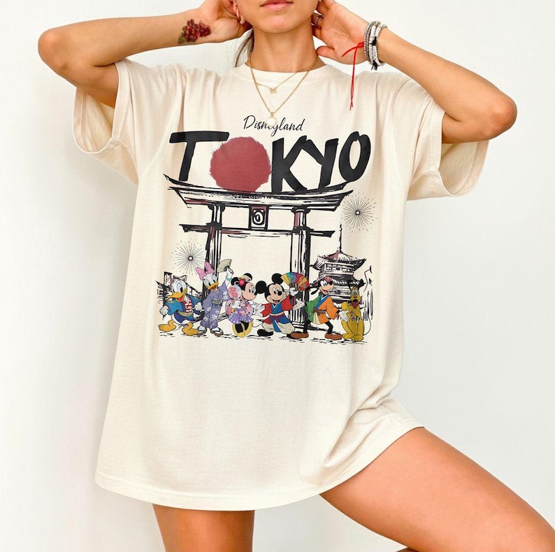 Puede incluir: Camiseta color crema con un dise&ntilde;o de Disneyland Tokyo. El dise&ntilde;o incluye la palabra "Tokyo" en negrita, un c&iacute;rculo rojo y una representaci&oacute;n de personajes de Disney frente a una puerta y una pagoda japonesas.