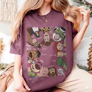K&ouml;nnte beinhalten: Burgunderfarbenes T-Shirt mit einer farbenfrohen Grafik von Toy Story-Charakteren. Das Design umfasst Woody, Buzz Lightyear, Jessie und andere Charaktere. Die Namen der Charaktere sind in einer handschriftlichen Schrift geschrieben.