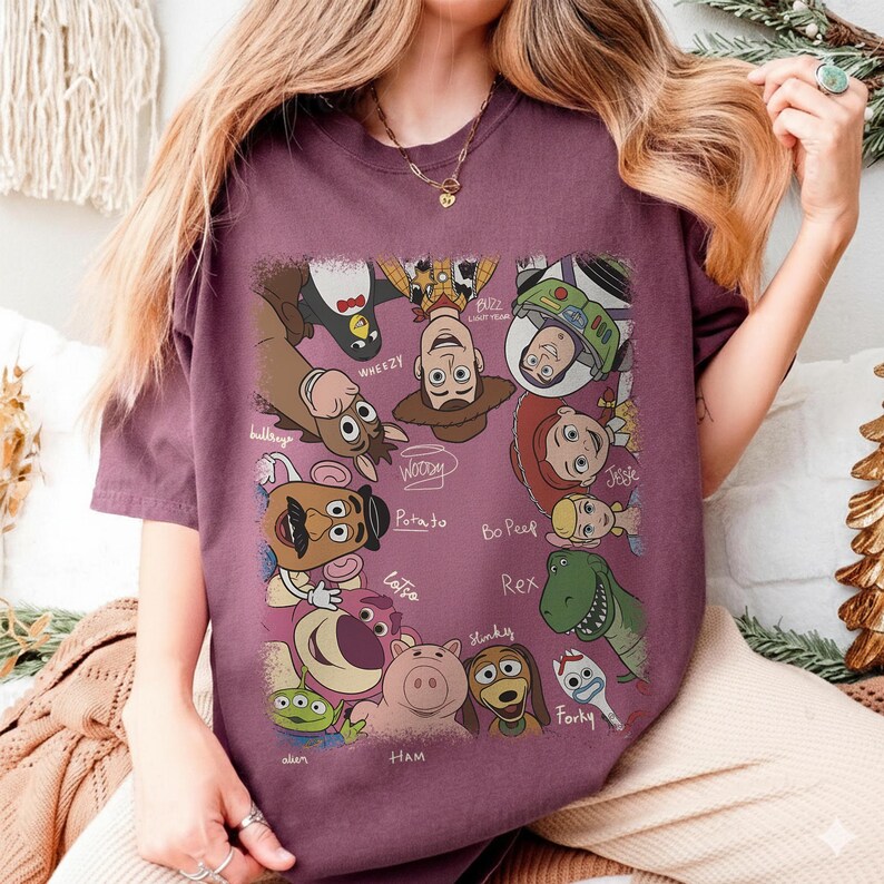 K&ouml;nnte beinhalten: Burgunderfarbenes T-Shirt mit einer farbenfrohen Grafik von Toy Story-Charakteren. Das Design umfasst Woody, Buzz Lightyear, Jessie und andere Charaktere. Die Namen der Charaktere sind in einer handschriftlichen Schrift geschrieben.