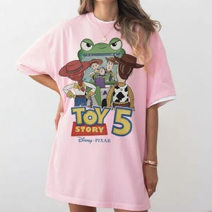 K&ouml;nnte beinhalten: Rosa T-Shirt mit einem Aufdruck von Toy Story-Charakteren und dem Text "Toy Story 5" mit dem Disney Pixar-Logo. Das T-Shirt hat kurze &Auml;rmel und einen wei&szlig;en Besatz. Das Design zeigt Woody, Buzz und andere Charaktere.