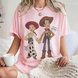 Może przedstawiać: Jasnor&oacute;żowa koszulka z kresk&oacute;wkową ilustracją Jessie i Woody z Toy Story trzymających się za ręce. Jessie ma na sobie czerwony kapelusz, białą koszulę i spodnie w krowie łaty. Woody ma brązowy kapelusz, ż&oacute;łtą koszulę i niebieskie spodnie. Na koszulce napisano "Woody".