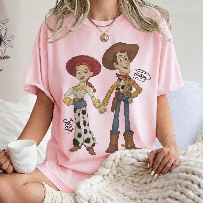 Może przedstawiać: Jasnor&oacute;żowa koszulka z kresk&oacute;wkową ilustracją Jessie i Woody z Toy Story trzymających się za ręce. Jessie ma na sobie czerwony kapelusz, białą koszulę i spodnie w krowie łaty. Woody ma brązowy kapelusz, ż&oacute;łtą koszulę i niebieskie spodnie. Na koszulce napisano "Woody".