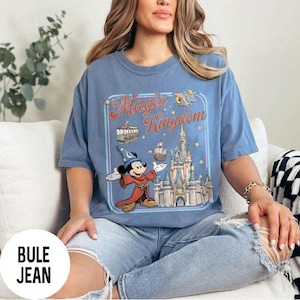 May include: Blue t-shirt with a graphic of Mickey Mouse as a sorcerer in front of a castle, with the words "Magic Kingdom" in a vintage style. The shirt is a light blue color.