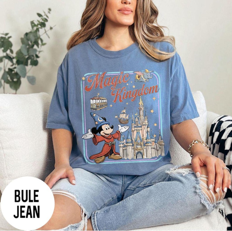 May include: Blue t-shirt with a graphic of Mickey Mouse as a sorcerer in front of a castle, with the words "Magic Kingdom" in a vintage style. The shirt is a light blue color.