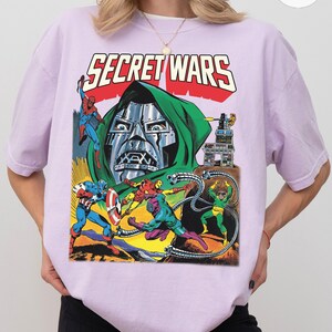 May include: A light purple t-shirt featuring a comic book illustration with the text "SECRET WARS". The graphic depicts various superheroes and villains in action, with a large metal-masked figure in the center.