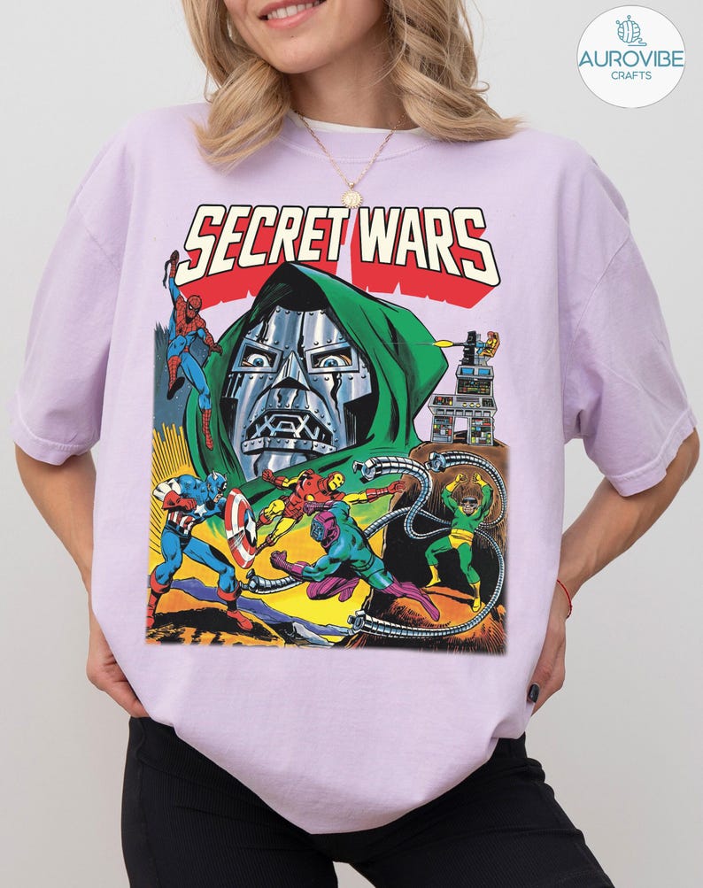 May include: A light purple t-shirt featuring a comic book illustration with the text "SECRET WARS". The graphic depicts various superheroes and villains in action, with a large metal-masked figure in the center.