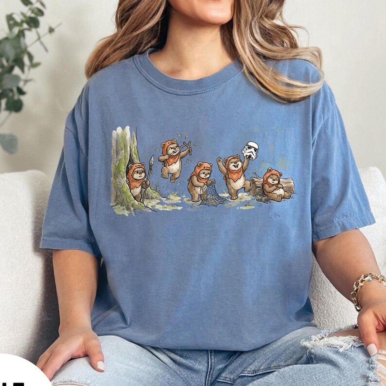 May include: A blue t-shirt featuring a cartoon illustration of Ewoks in a forest setting. The Ewoks are depicted in various poses, including holding spears, a net, and a Stormtrooper helmet. The shirt has a crew neck and short sleeves.