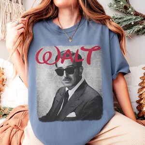 May include: A blue t-shirt featuring a black and white image of a man wearing sunglasses and a suit. The word "WALT" is written in red cursive above the image. The shirt has short sleeves.